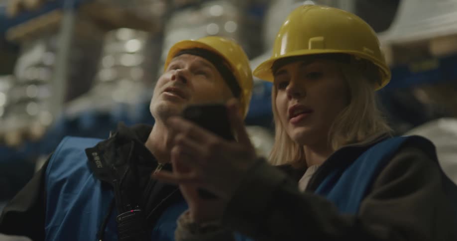 A man and a woman with vests and hardhats in a warehouse with shelves in the background