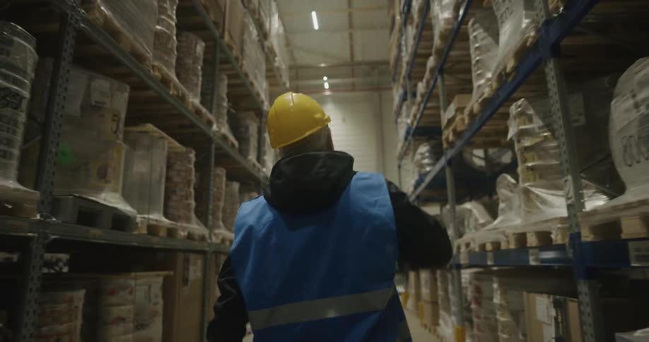 A man with a walkie talkie a hardhat and a vest walking in a warehouse with shelves