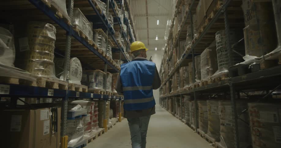 A man with a hardhat and a walkie talkie in a warehouse with shelves