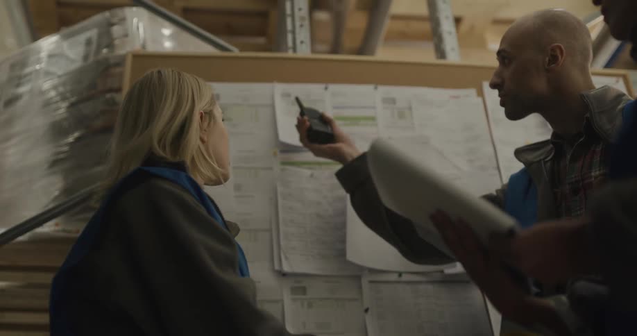 A group of coworkers speak and examine a clipboard while standing in front of a bulletin board in a warehouse