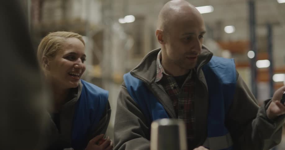 A man with a vest using a smart phone to show a video to a woman in a warehouse