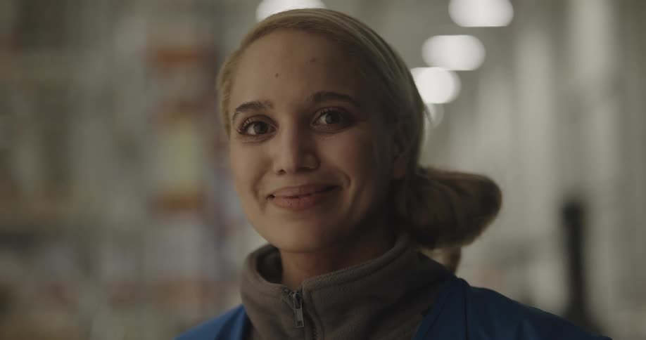 A woman with a vest in a warehouse standing and smiling with shelves and lights in the background