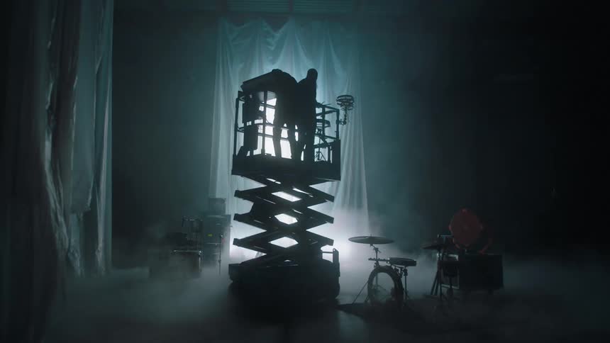 The silhouette of two musicians rising up on a scissor lift with fabric hanging and fog and lights creating a dramatic set