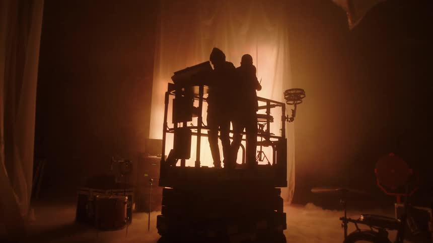 A silhouette of two people on a stage on a lift with one playing the drums and the other one playing a keyboard