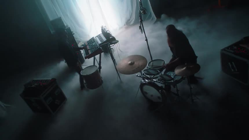 Two men on a stage with smoke with one playing the drums and the other one playing a drum and a keyboard