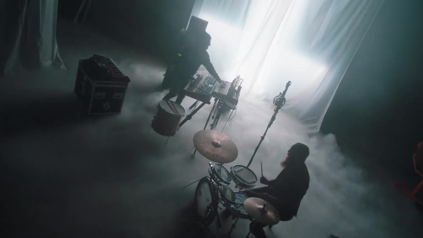 A band on a foggy stage playing alone in front of a curtain with a strobing light behind it while smoke pours in from underneath