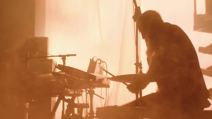A keyboardist and drummer play their instruments as fog swirls around them in a dimly lit room