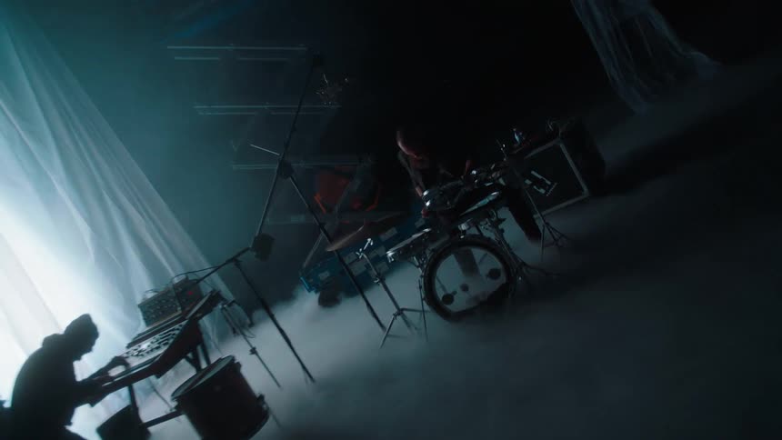 A drummer and keyboardist play their instruments on a dimly lit stand with its floor shrouded in fog