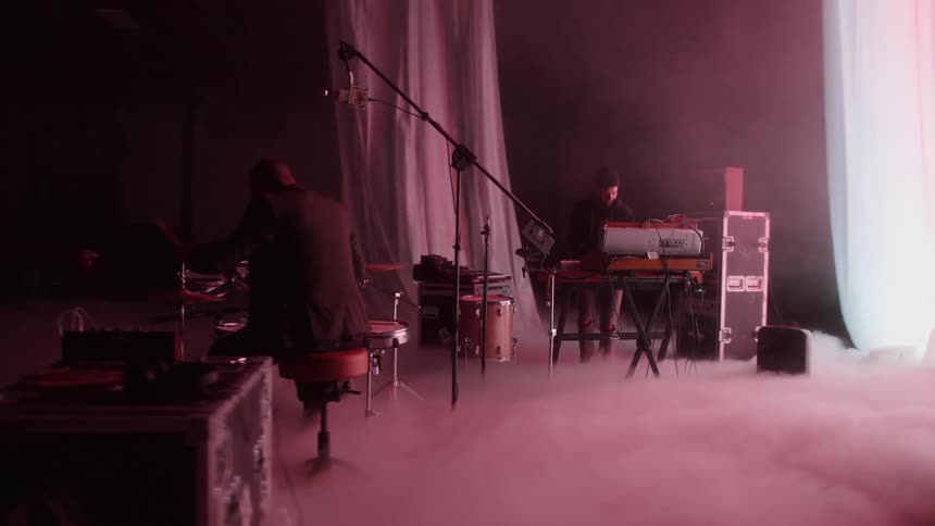 Two men play musical instruments as lights flash and fog moves across the floor in a video set
