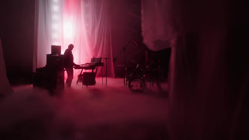Two silhouettes of two men where one is playing a drum set while the other dance and plays an electric keyboard on a stage with red and white lights flashing behind a curtain