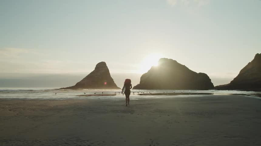 Sun shines past rock outcroppings on beach to silhouette female surfer carrying surfboard toward ocean