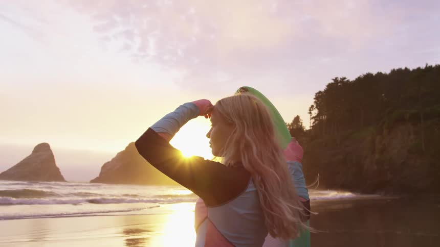 Young blonde woman wearing a colorful wet suit holding a green surfboard on the coast of a beach at sunset