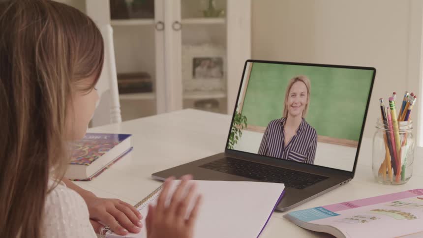 A girl waves at her teacher who is on screen on a laptop