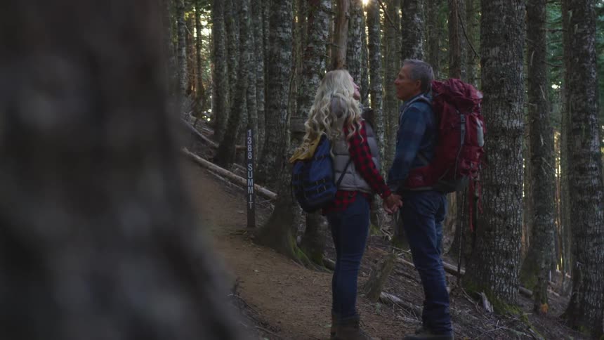 A man and a woman with backpacks holding hands in a forest with trees