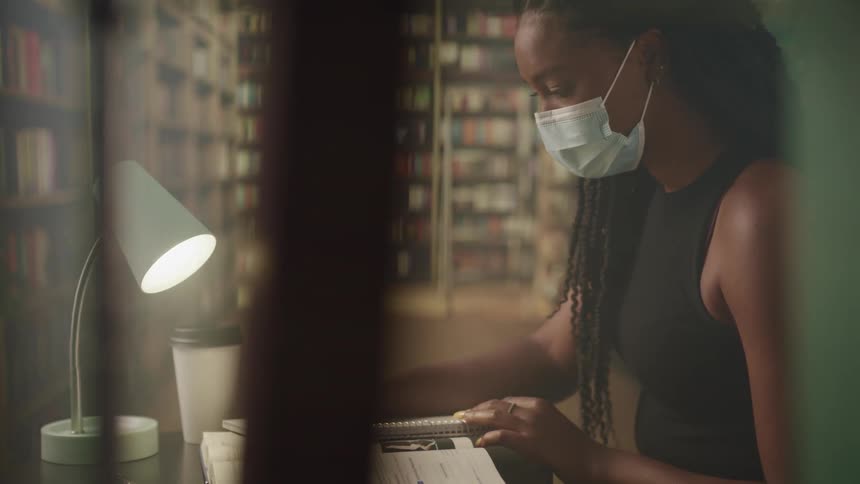 Woman with braids and a mask writes in a notebook in the library with bookshelves in the background