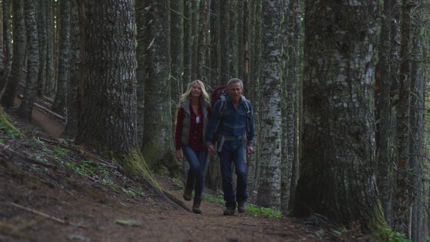 A man and woman hold hands as they walk together along a dirt trail through a dense forest full of tall trees