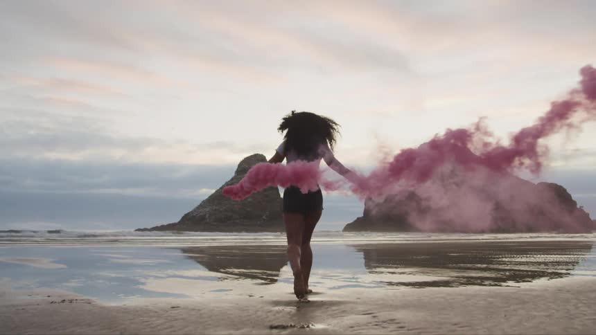 A woman skips on the beach holding pink smoke flares
