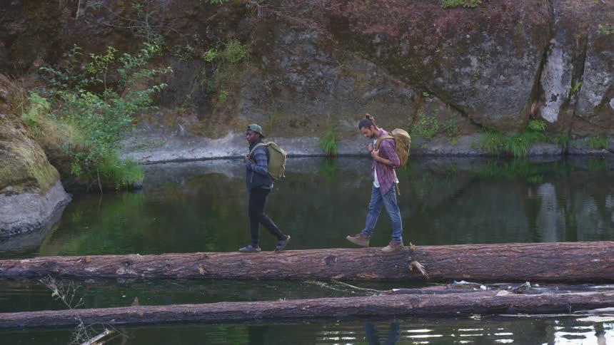 Two men carrying backpacks walking over a fallen tree trunk in the surface of a lake in the morning