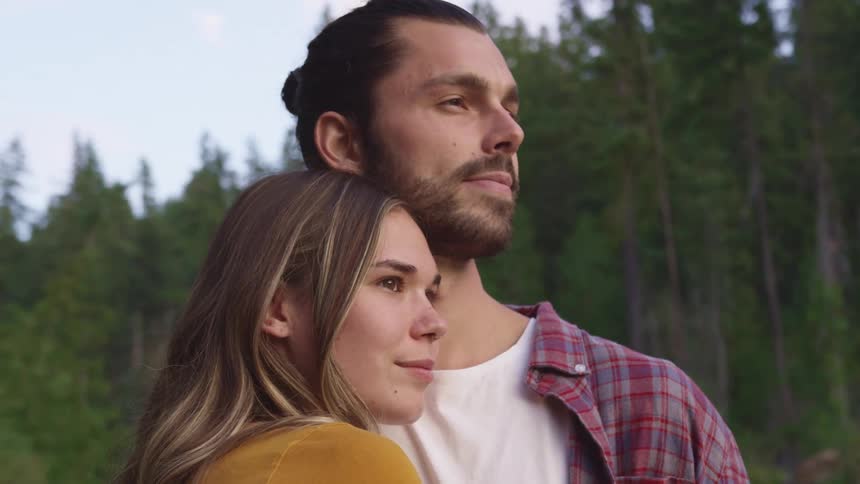 A man and woman embrace while standing together in a woodland during the day