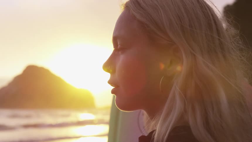 Woman standing on beach near surfboard is lit by sun sitting on horizon