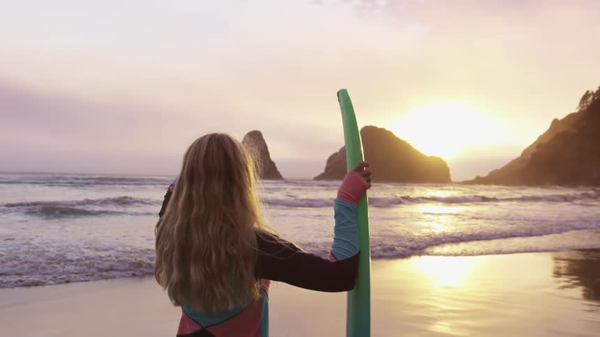 A female stands on the beach by the ocean water holding a surfboard and staring at the low sunlight