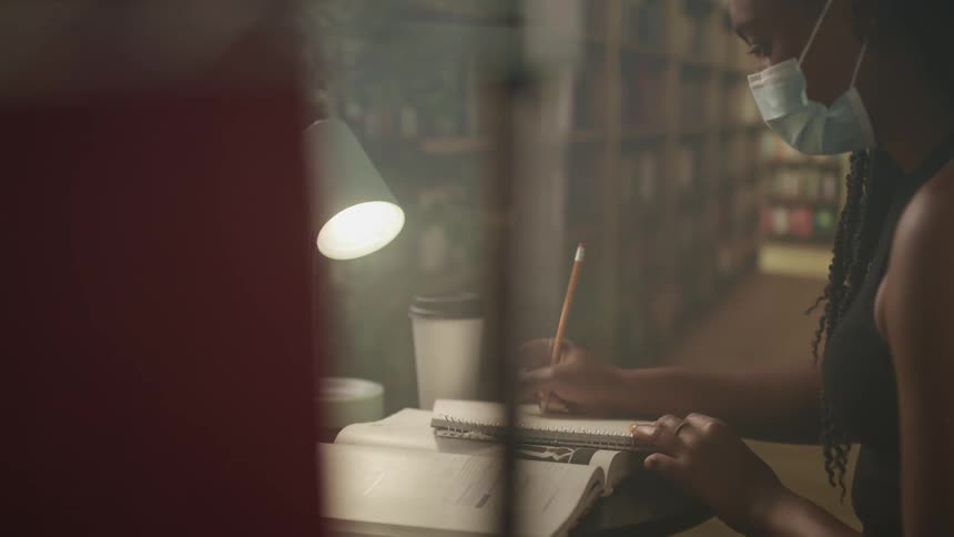 Woman with braids a mask and a black sleeveless shirt writes down in the library with bookshelves in the background