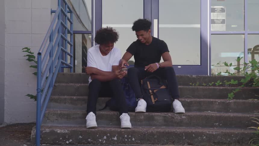 Two black males sitting on building stairs looking at a phone and laughing