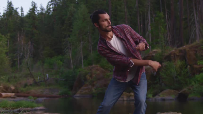 A white male stands on the edge of the river and tosses a stone to skip on the waters surface