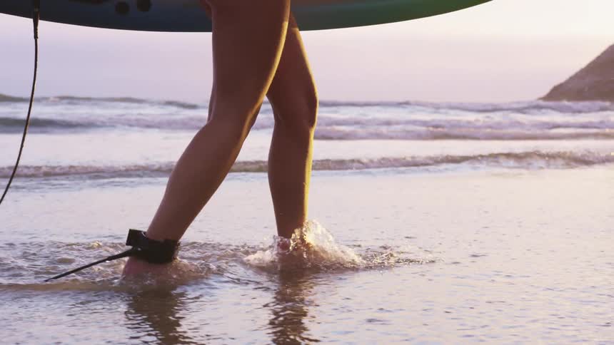A woman walking on a beach holding a surfboard with the ocean sky and clouds in the background