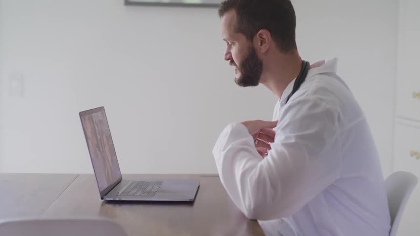 A male doctor sits at a table having a video call on a laptop
