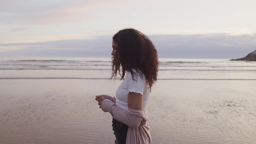 A young woman stands alone at the beach near the to where the waves gently wash ashore