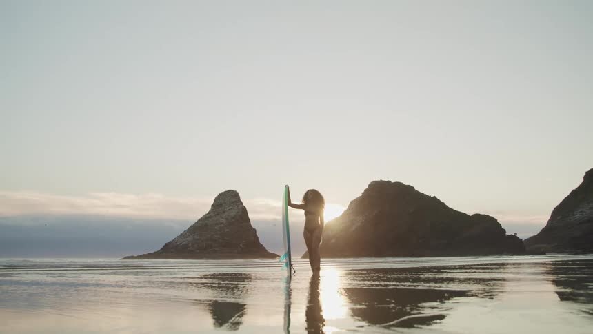 A woman stands in the low tide on a beach at sunset while holding a surfboard
