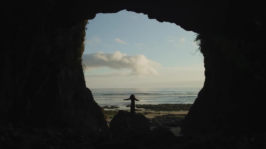 Woman wearing a black dress standing on the entrace of a cove near the beach at sunset