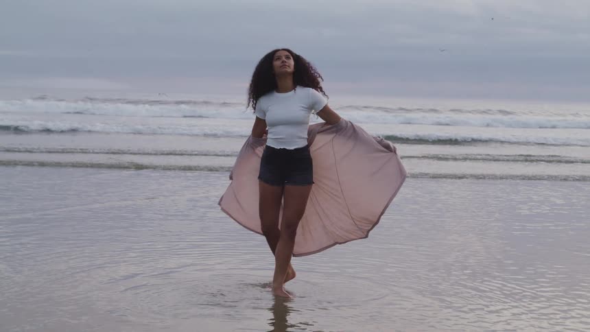 A young woman stands in the shallow ocean water on the beach spinning with a shaw behind her blowing in the wind