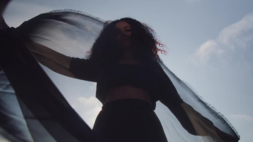 Woman wearing a black dress waves a black see through cloth in the air at sunset