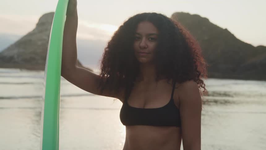 A black female standing on the beach holding a surfboard