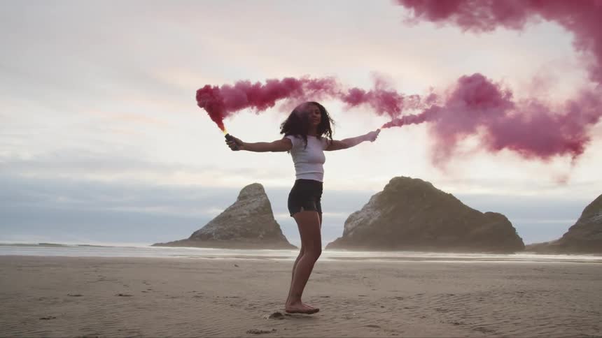 Woman walks along beach toward water slowly turning and spreading purple smoke fromhandheld burning cylinders