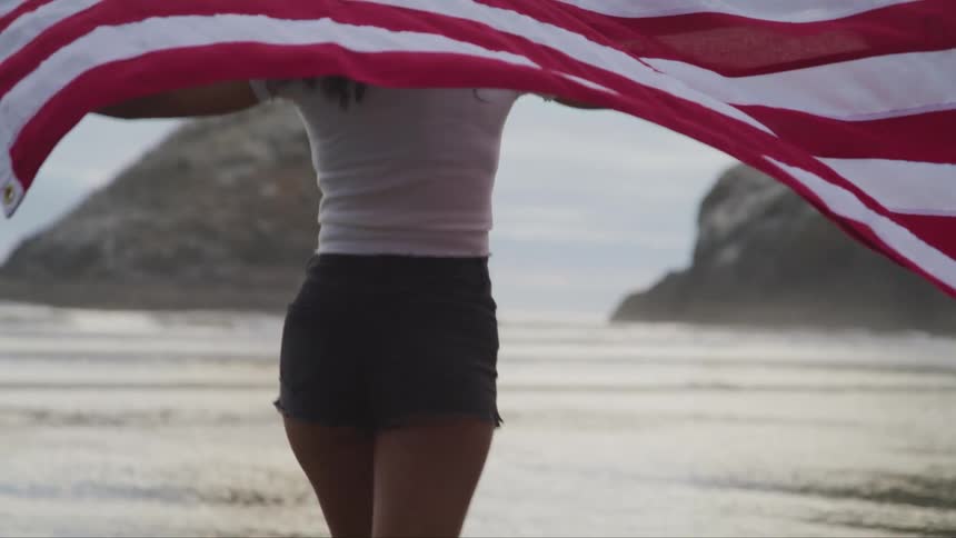 A barefoot woman skipping on a sandy beach with an american flag trailing behind her