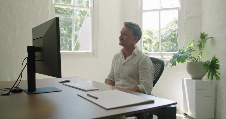 A middle aged man waves and blows kisses toward a computer monitor while video chatting at a desk during the day