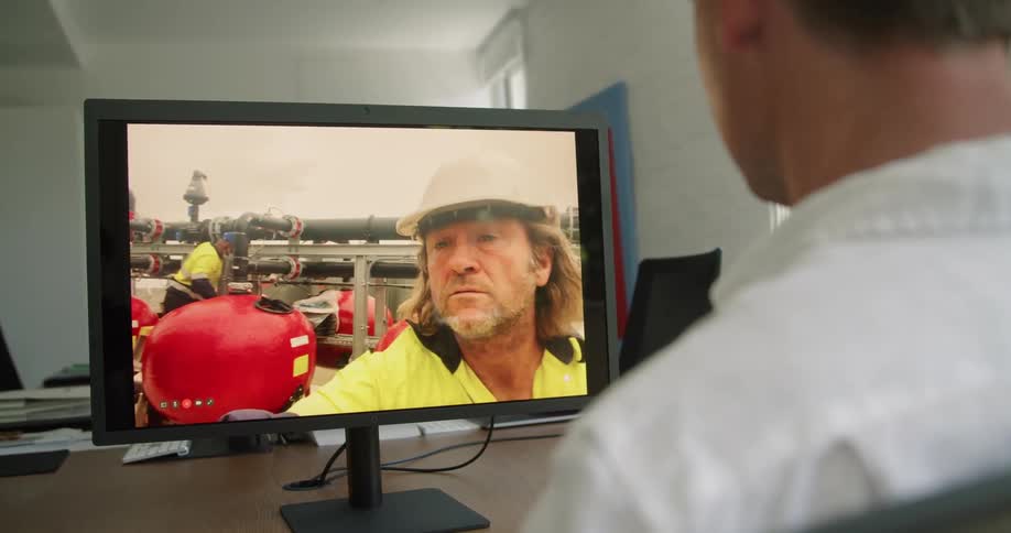 A man sitting on a chair at a desk in front of a monitor with a wall and window in the background