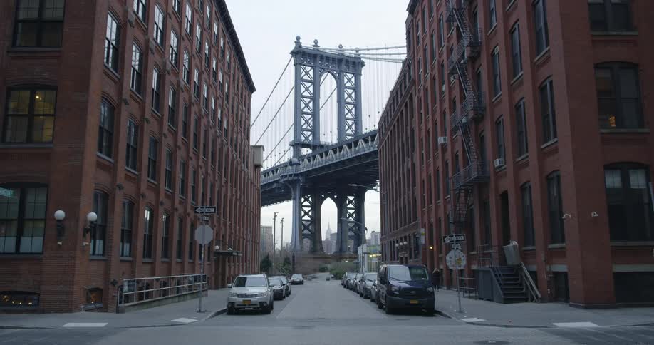 A person crossing a city street with two buildings and a bridge in the background