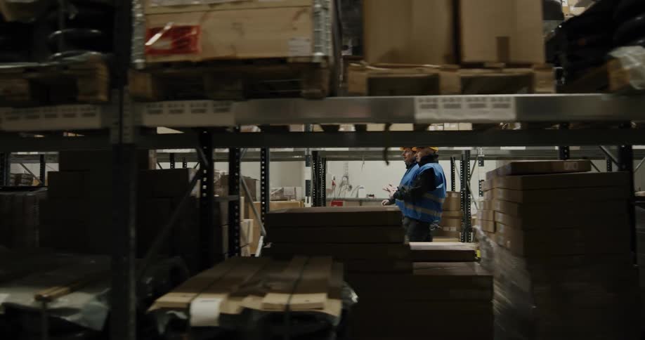 Two men in vests and brightly colored hard hats walk between large shelves in a warehouse