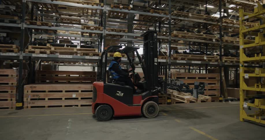 A man drives a forklift through a narrow aisle in a warehouse during the day