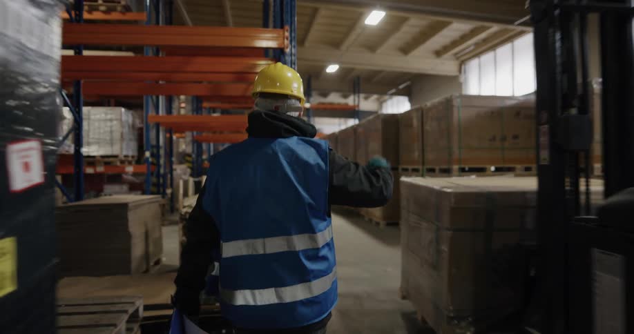 A man in personal protective equipment waves at a coworker operating a forklift as he walks through a warehouse