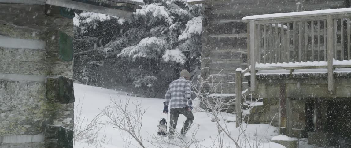 A man approaches a log cabin in a snowstorm