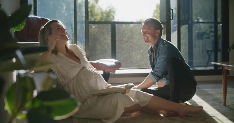A man sitting on the floor at home drinks his coffee and reaches over and touches his wifes pregnant belly
