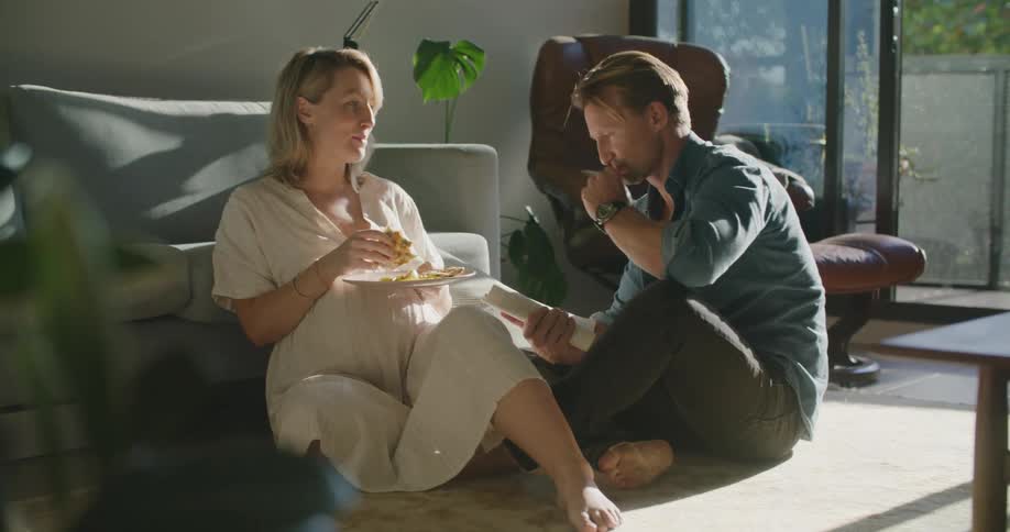 Pregnant couple sitting in the livingroom at home talking and eating