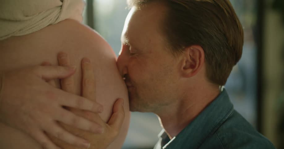 Kneeling man listens to and kisses womans pregnant belly