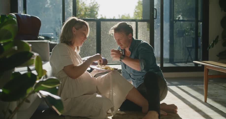Young pregnant couple sitting in the living room eating food at sunset