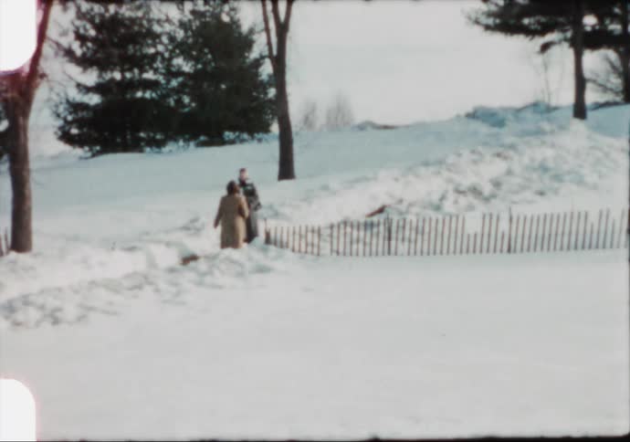Two people walking on snow next to a fence with trees the sky and clouds in the background